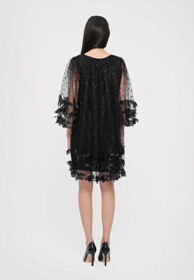 Crna, slojevita haljina s prozirnom tkaninom s cvjetnim uzorcima, rukavima u obliku leptira i naborima na rubu. Crne cipele s visokom petom.