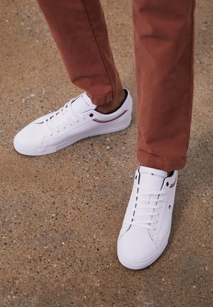 Personne portant des baskets blanches Tommy Hilfiger et un pantalon couleur rouille debout sur une surface en béton tachetée.