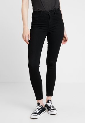 Vaqueros skinny negros de cintura alta con una tela elástica y elegante. Cuenta con cinco bolsillos y un cierre de botón clásico. Estilizados con zapatillas negras.