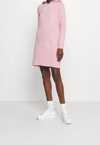 Vestido de sudadera rosa hecho de una tela suave, con un bolsillo delantero y un logo. Zapatillas blancas completan el atuendo, proporcionando un aspecto casual.