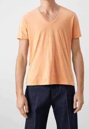 Man draagt een lichtoranje T-shirt met V-hals en korte mouwen en donkerblauwe spijkerbroek tegen een egale achtergrond.
