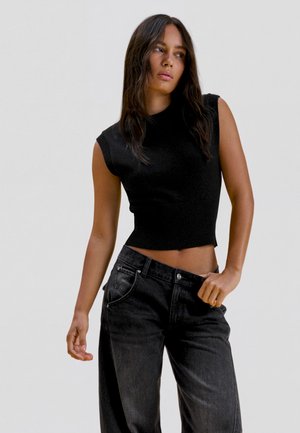 Vrouw met lang donker haar die een mouwloos zwart crop top en losse zwarte spijkerbroek draagt, staand tegen een effen lichte achtergrond.