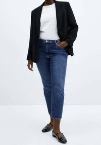 Blazer noir sur un t-shirt blanc, associé à un jean denim bleu et des chaussures plates noires. Le jean a une coupe droite et une légère décoloration.