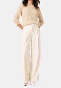 Pull en tricot beige à manches trois-quarts associé à un pantalon large blanc taille haute ; texture lisse et lignes épurées.