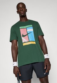 Zaļš kokvilnas T-krekls ar krāsainu grafiku, kas ietver rozā, zilu un dzeltenu sekciju, kā arī melnu Nike logo. Modelis arī nēsā rokassprādzes.