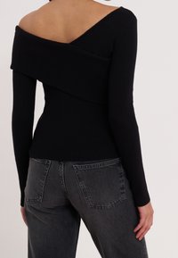 Frau in einem schwarzen, schulterfreien Rippstrickpullover, kombiniert mit hoch taillierten dunkelgrauen Jeans, von hinten gezeigt.