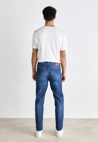 Biała męska koszulka z krótkim rękawem w połączeniu z niebieskimi dżinsami denimowymi, posiadającymi pięć kieszeni i czerwoną naszywkę z logo na pasku.