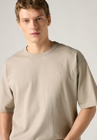 Ανοιχτό ταουπ κοντομάνικο μπλουζάκι από απαλό βαμβακερό ύφασμα, με στρογγυλή λαιμόκοψη και μικρό, μινιμαλιστικό λογότυπο στο στήθος.