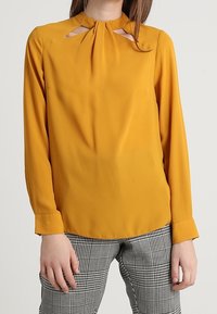 Senapsgul blus med hög halsringning, nyckelhålsutskärningar, långa ärmar och en mjuk textur. Matchad med rutiga grå byxor.