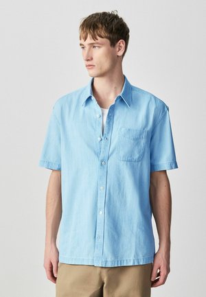 Muž v svetlomodrej krátkej košeli na gombíky cez biele tričko a béžových nohaviciach, pozerajúci sa doľava na neutrálnom pozadí.