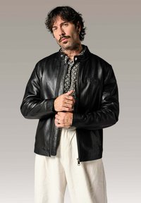 Veste en cuir noir avec fermeture éclair et col côtelé, ornée d'un logo en relief. Portée par-dessus une chemise à motifs ; le modèle se tient devant un fond neutre.