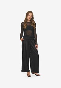 Ženska oblečena v prosojno črno majico z dolgimi rokavi in malimi pikami, široke črne hlače in črne sandale na peto, stoji z eno roko v žepu.