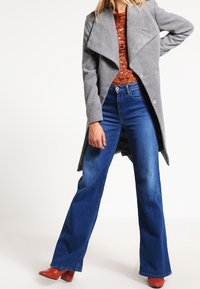 Abrigo de lana gris con amplias solapas, llevado sobre una blusa de textura color óxido y vaqueros de campana azules, complementado con botas de cuero rojas de tacón.