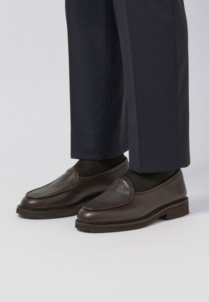 Mocasines de cuero marrón con acabado texturizado, punta redonda y suela de perfil bajo, combinados con pantalones oscuros y calcetines negros.