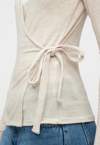 Beige pitkähihaine paita, jossa on kietoutumismalli ja solmittu yksityiskohta takana. Pehmeä kangas ja hienovarainen rakenne. Yhdistetty farkkujen kanssa.