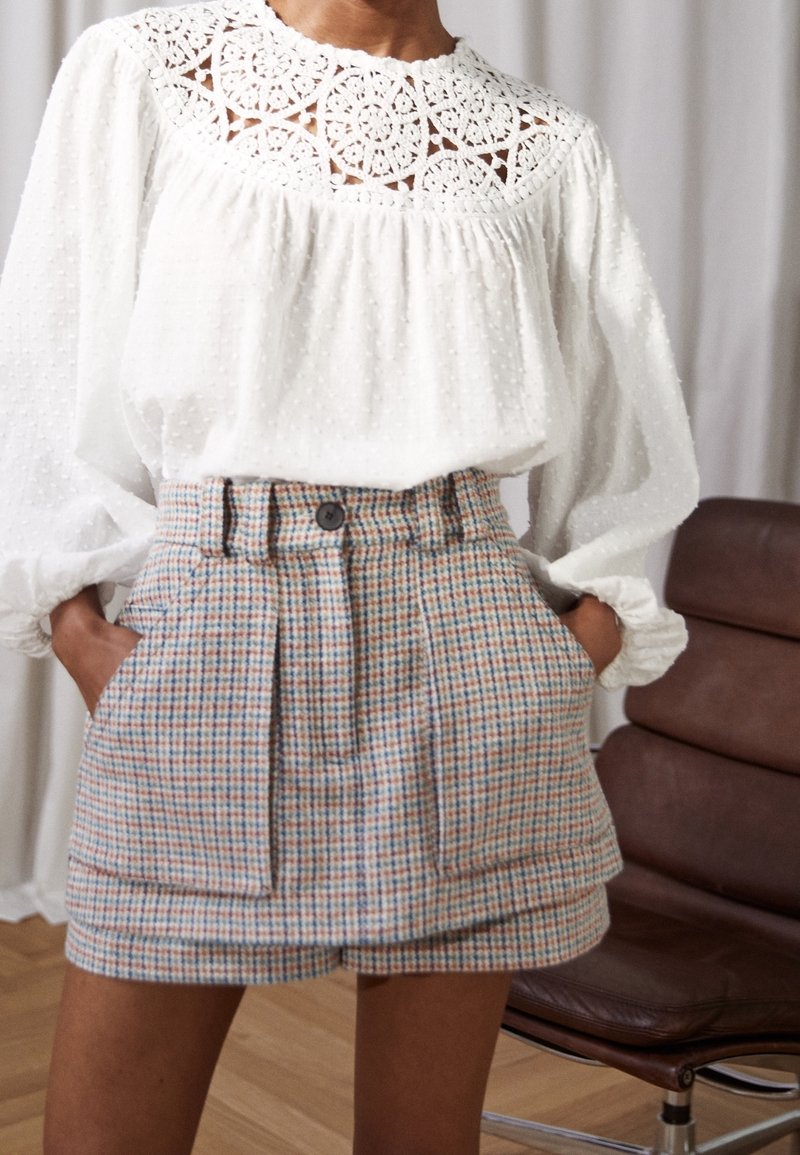 Hvid bluse med tekstureret stof og blonde detaljer ved udskæringen, parret med ternede højtsiddende shorts i lys blå, beige og orange.