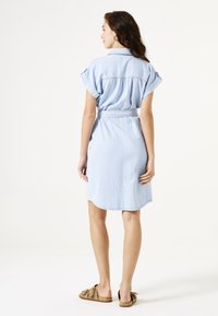 Helles blaues Jeanshemdkleid mit kurzen, umgeschlagenen Ärmeln, tailliertem Design und abgerundetem Saum. Getragen mit beigen Slip-On-Schuhen. Glatte Textur.
