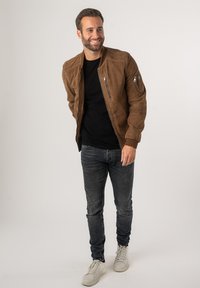 Giubbotto bomber in suede marrone con polsini a costine, tasche con zip, indossato su una maglietta nera, abbinato a jeans scuri e sneakers bianche.