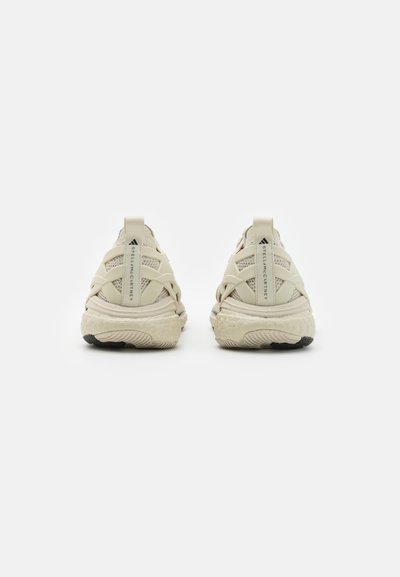 Beige athletic shoes with a mesh upper, crisscrossed straps, and a textured sole. Features a logo on the heel and a rounded toe design.
