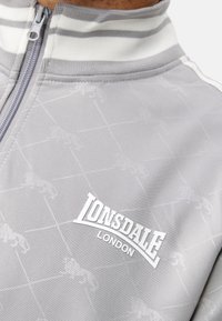 Grijze ritsjack met een subtiel leeuwpatroon, wit logo "Lonsdale London" en een gestreepte kraag. Gemaakt van glad synthetisch materiaal.