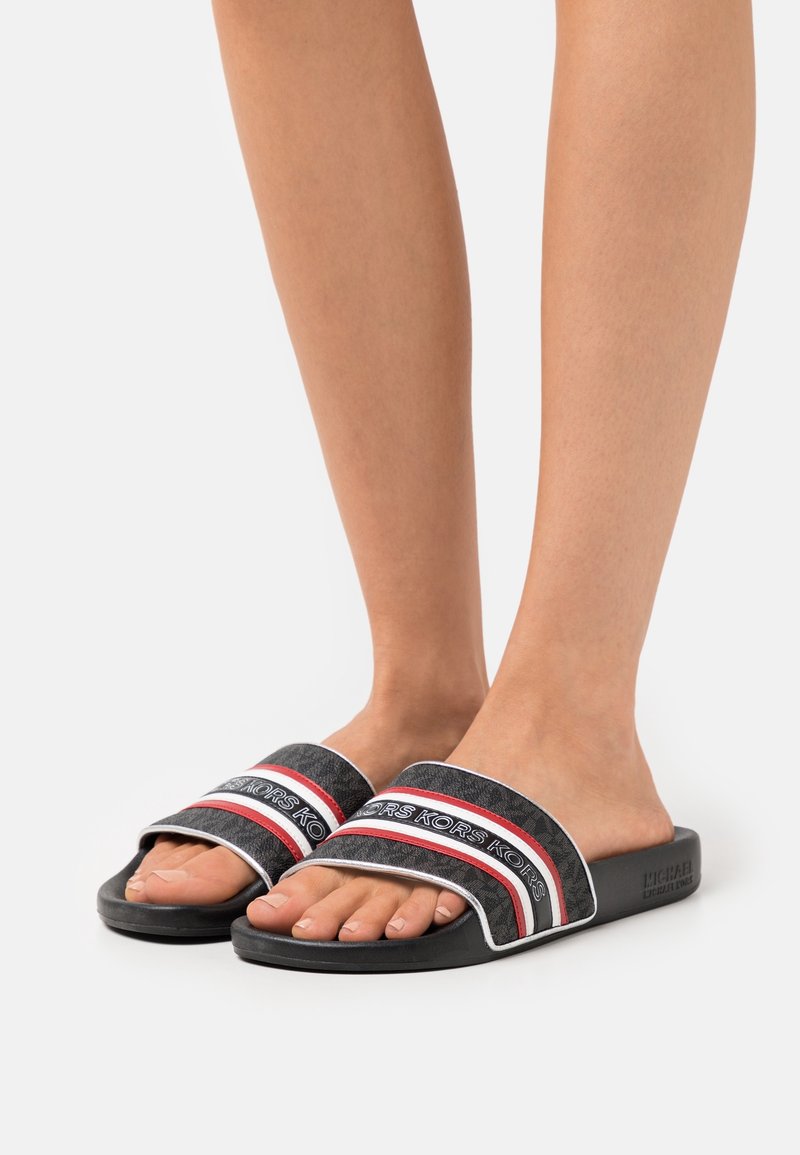 Pieds portant des sandales noires à enfiler avec des brides rayées rouges et blanches marquées du logo « KORS » sur un fond clair uni.