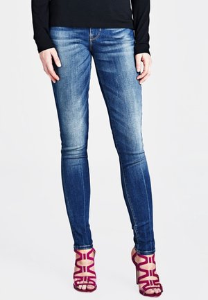 Personne portant un jean skinny bleu délavé et des sandales à talons hauts magenta avec des lanières, debout devant un fond blanc uni.