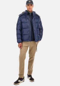 Marinblå pufferjacka med huva, sidfickor och en glansig yta, tillsammans med beige byxor och svarta sneakers med flerfärgade accenter.