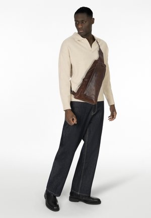 Braune Leder-Schultertasche mit einem eleganten Design, ausgestattet mit einem diagonalen Reißverschluss und einem verstellbaren Tragegurt, getragen über einem beigen Pullover und dunklen Jeans.