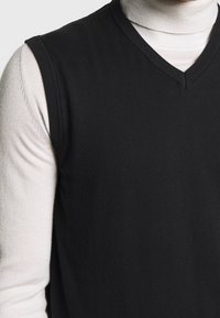 Black sleeveless V-neck sweater worn over a light beige long-sleeve turtleneck.