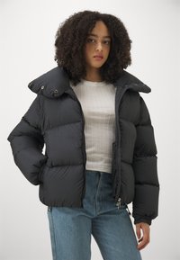 Fekete puffermellény magas, túlméretezett gallérral, cipzáras előlappal, rugalmas mandzsettákkal és oldalsó zsebekkel. Simára kidolgozott textúrával, hangsúlyos párnázottsággal.