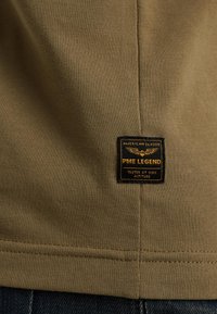 Beiger Stoff mit einem schwarzen und goldenen rechteckigen PME Legend-Patch, auf dem "American Classic, Tested at High Altitude" in der Nähe einer Naht steht.