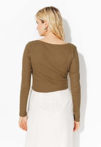 Haut marron à manches longues avec un décolleté festonné, une texture côtelée et une coupe ajustée, associé à une jupe plissée blanche. Vue de dos.