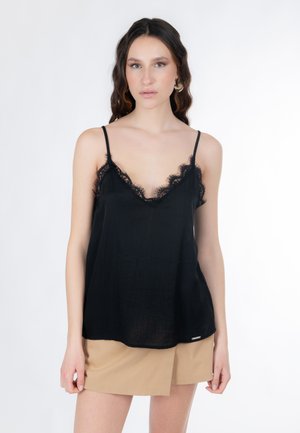 Femme aux cheveux ondulés portant un haut camisole orné de dentelle noire et une jupe portefeuille beige sur un fond blanc uni.