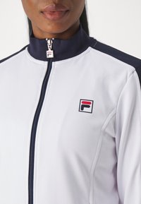 Nærbillede af en person iført en hvid lynlåssweatshirt med navyblå krave, navyblå skulderstriber og et lille FILA-logo på brystet.