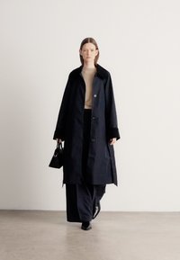 Hosszú tengerészkék kabát nagy gallérral, gombos záródással és zsebekkel, bézs garbóval és sötét nadrággal párosítva. A modell egy fekete kézitáskát tart.