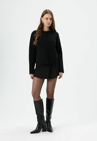 Schwarzer Strickpullover mit Rundhalsausschnitt, kombiniert mit einem schwarzen plissierten Minirock. Abgerundet wird das Outfit durch kniehohe schwarze Lederstiefel und transparente Strumpfhosen.