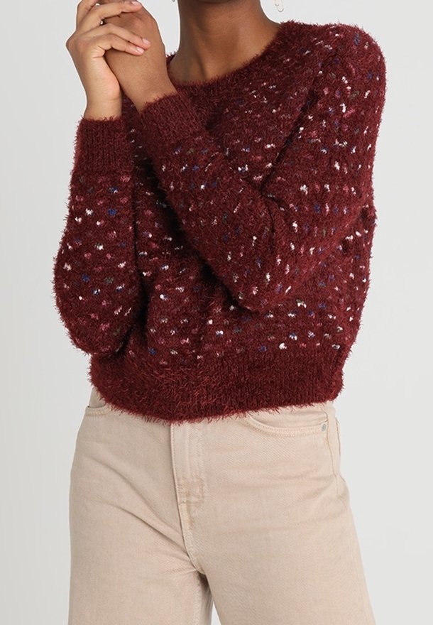 Maglione borgogna morbido con macchie multicolori, scollatura tonda e maniche lunghe, abbinato a jeans beige chiaro.