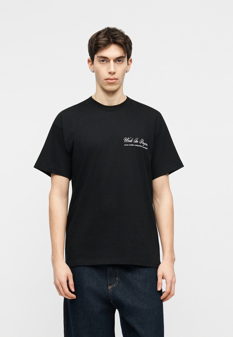 Giovane uomo con capelli corti e scuri, che indossa una T-shirt nera con testo bianco sul petto sinistro e jeans blu scuro, in piedi contro uno sfondo neutro.