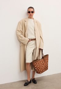 Kvinna i beige lång kappa, krämfärgad tröja och shorts, svarta loafers, solglasögon, håller en stor brun prickig toteväska, står mot en vit vägg.