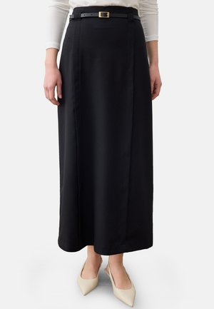 Femme portant une longue jupe noire avec une fine ceinture noire et des chaussures beige à bout pointu et bride arrière, debout avec les mains détendues le long du corps.