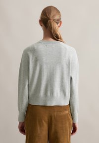 Grå stickad tröja med rund halsringning, ribbad nederkant och långa ärmar; subtil textur och åtsittande design. Åtföljs av bruna kordfläskbyxor.