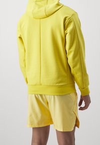 Sweat à capuche jaune à coupe décontractée, avec une capuche et des poignets côtelés. Associé à un short jaune assorti avec des fentes latérales et une texture lisse.