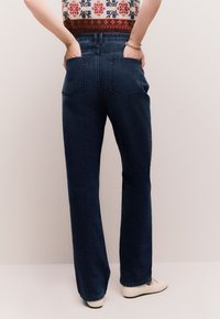 Tamsiai mėlyni džinsai iš džinso audinio, platėjančiu siluetu, aukštu liemeniu ir užpakalinėmis kišenėmis. Kelnaičių apačia šiek tiek pjūkliška, derinama su baltais batais.