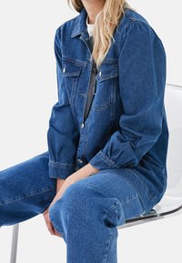 Vaaleansininen farkkutakki, jossa on pitkät hihat, kaksi rintataskua ja napitettu suljenta, yhdistettynä vastaaviin farkkuhousuihin. Sileä pinta.