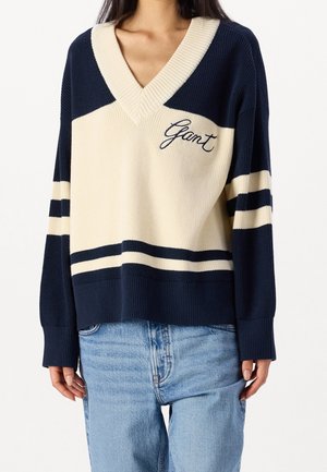 Femme portant un pull en tricot bleu et crème à col en V avec broderie "Gant" et rayures bleues, associé à un jean bleu clair.