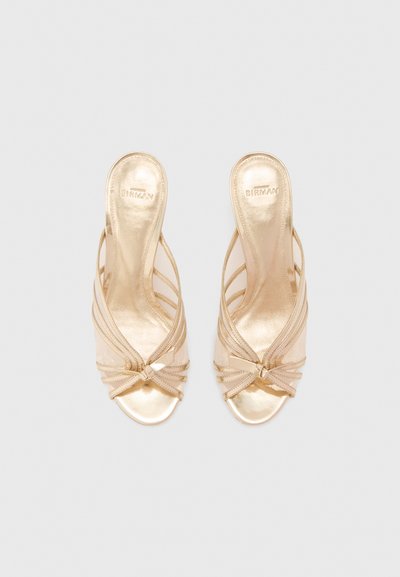 Ζευγάρι από ανοιχτές χρυσές σανδάλες με ψηλό τακούνι, διασταυρωμένους ιμάντες και λεπτομέρεια φιόγκου, σε άποψη από πάνω πάνω σε ανοιχτό γκρι φόντο.