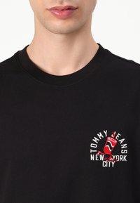Black cotton t-shirt featuring a round neckline and a colorful embroidered logo with a drink cup and text "TOMMY JEANS NEW YORK CITY."