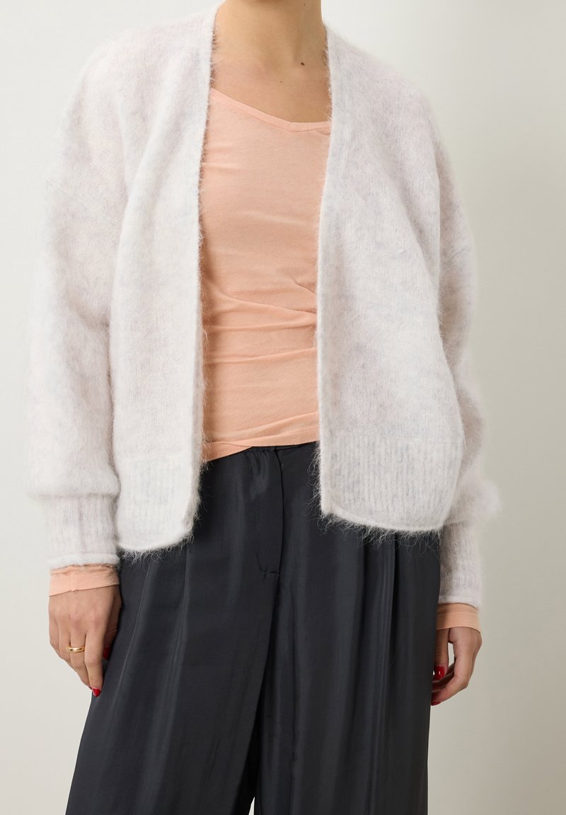 Valkoinen, pörröinen neuletakki, jossa on avonainen etuosa, pitkät hihat ja ribbausvarsi. Päällä vaaleanpunainen kapea toppi ja löysät mustat housut.