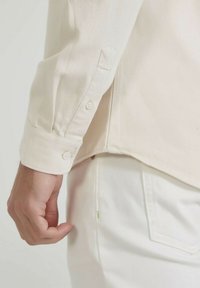 Gros plan sur le bras gauche d'une personne portant une chemise à manches longues crème avec des poignets boutonnés et un pantalon blanc cassé avec une poche latérale visible.