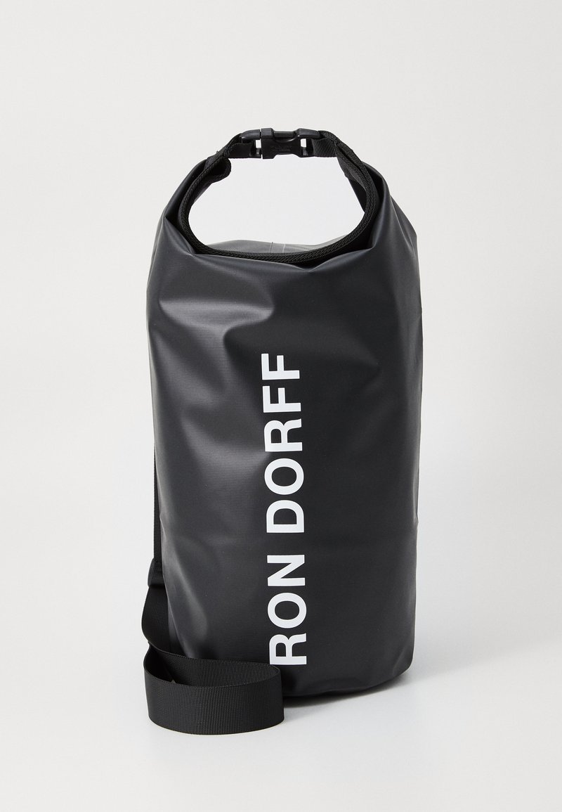 Must mustest tõmblukuga kott, mis on valmistatud vastupidavast materjalist, omades õlavarre rihma ja esiküljel valget logo teksti. Sile tekstuur.